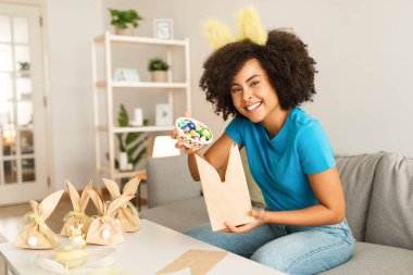 Güzel siyahi kadın Paskalya ziyafeti veriyor, sevimli tavşan kulaklı mutlu Afrikalı kadın saç bandı takıyor, çikolatalı yumurtaları ev yapımı kağıt torbaya koyuyor ve kameraya gülümsüyor.