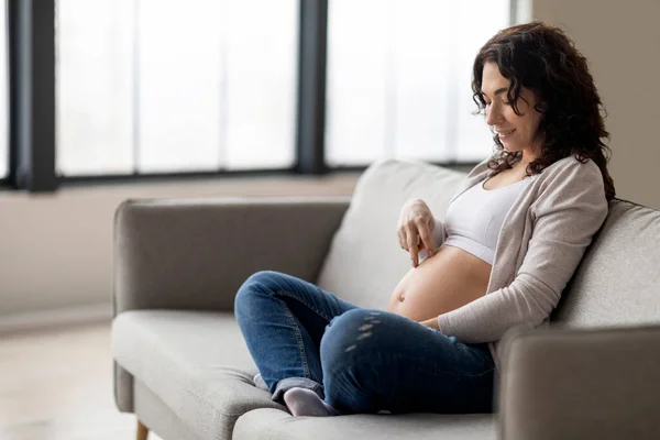 若い妊婦の肖像画彼女の大きな腹を指で歩いている間に自宅でソファに座って 妊婦のために準備を笑顔 彼女の妊娠時間を楽しんで スペースをコピー — ストック写真