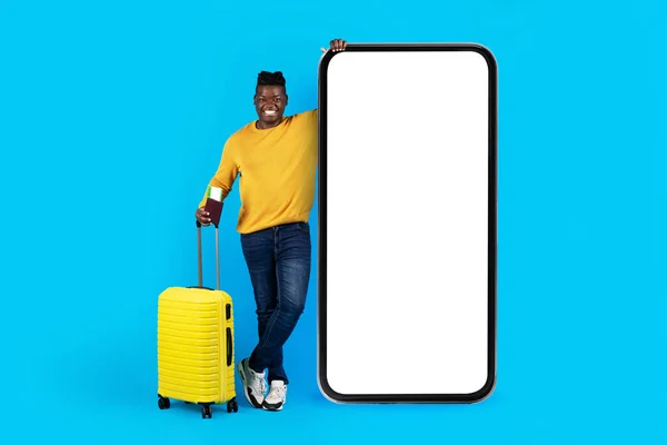 Travel App. Smiling Black Man With Suitcase Standing Near Big Blank Smartphone With White Screen, Happy Young African American Man Advertising Modern Application For Tickets Booking, Mockup