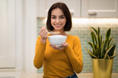 Olumlu, neşeli, oldukça ince bir bayan evde sağlıklı bir kahvaltı yapıyor, elinde yulaf ezmesi, tahıl gevreği ve kaşık ile gülümseyen bir kadın, mutfak içi, kopyalama alanı.