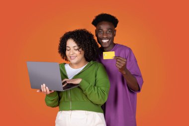 Mutlu neşeli melez ırk aşıkları genç siyah adam ve Latin tombul kadın birlikte alışveriş yapıyor ya da online bankacılık yapıyor, dizüstü bilgisayar ve kredi kartı kullanıyor, kırmızı stüdyo arka planı, kopyalama alanı kullanıyor.