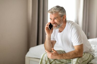 Neşeli gülümseyen, çekici, orta yaşlı, beyaz saçlı, evde yatakta oturan, telefonla konuşan, reklam için fotokopi odasına bakan, pijama giyen bir adam.