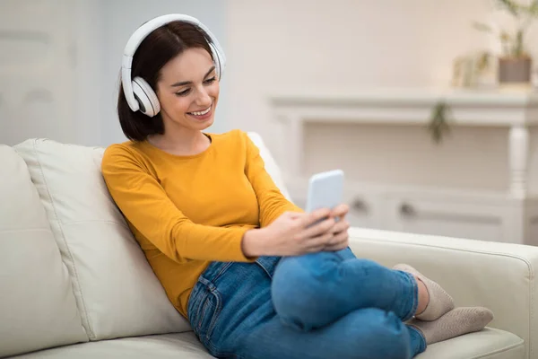 美しい幸せな陽気な若いブルネットの女性が一人でリビングルームでソファに座って 自宅で現代的なスマートフォンやワイヤレスヘッドフォンを使用して ビデオコンテンツをオンラインで視聴し スペースをコピー — ストック写真
