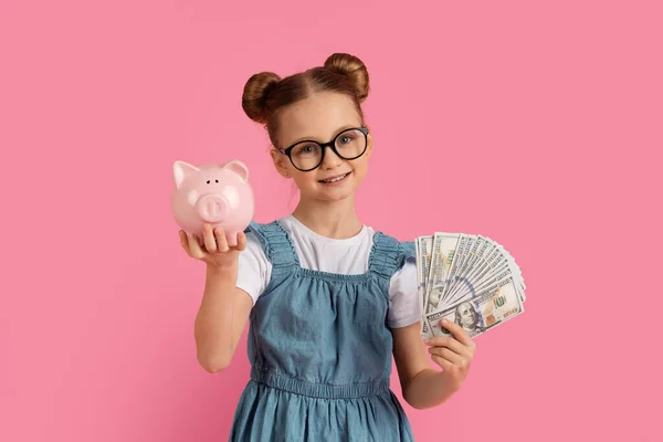 Money Saving. Cute Little Girl In Eyeglasses Holding Piggybank And Dollar Cash In Hands, Happy Female Child Enjoying Economy, Standing Isolated Over Pink background In Studio, Copy Space