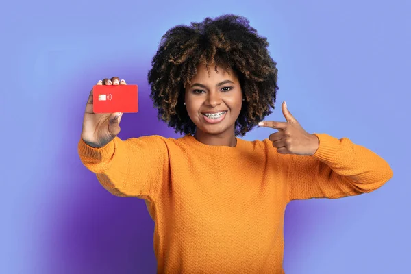 清凉开朗的微笑 美丽的年轻黑人女士在镜头前 指着手中的银行卡 紫色的工作室背景 无接触支付 现代银行业务概念 — 图库照片