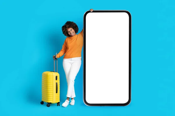 网上预订 预订移动应用程序 快乐而又时尚的年轻黑人女性 穿着休闲装 带着大大的手机摆出一副白色空白屏幕 蓝色背景 笑脸的样子 — 图库照片