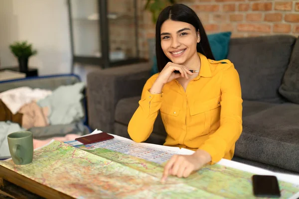 Ready to trip and vacation plan concept. Happy spanish lady sitting with map and pointing finger at desired location, looking and smiling at camera, planning holiday at home