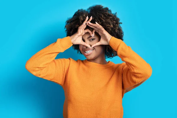 目と笑顔の横にハート型のジェスチャーを示すオレンジでかわいい巻きかなり若い黒人女性 女性は青いスタジオの背景の上にいちゃつく ジェスチャー 感情の概念 — ストック写真