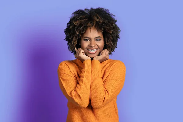 顔とカメラで笑顔の横に手を保持歯のブレースとオレンジでかわいい陽気なかなり若いアフリカ系アメリカ人女性 紫色のスタジオの背景に孤立 人間の感情やジェスチャーの概念 — ストック写真