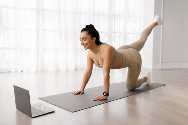 Çevrimiçi antrenman. Spor kıyafetleri içinde sportif, esmer, güzel bir kadın. Evde yoga minderi üzerinde egzersiz yapıyor. Bacaklarını esnetiyor, bilgisayar başında spor videosu izliyor ve gülümsüyor.