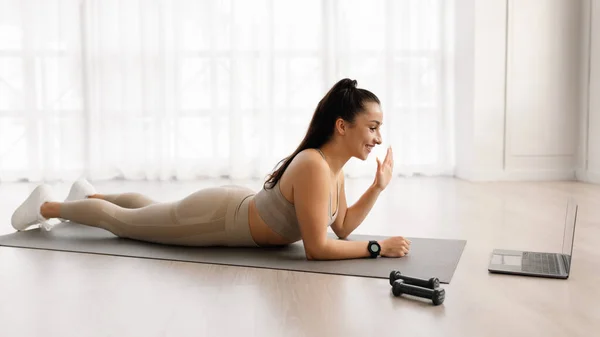 Fit young brunette long-haired woman in sportswear doing yoga, lying on fitness mat and waving at laptop screen, training with remote fitness coach, living room interior, side view, copy space