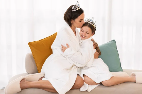 Mom And Daughter Wearing White Bathrobes And Toy Crowns Hugging At Home, Portrait Of Young Mother And Cute Female Child Resting On Couch After Beauty Spa Day, Bonding Together, Copy Space