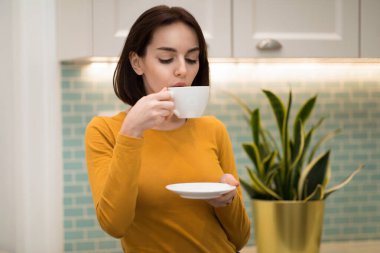 Ev kıyafetleri içinde güzel esmer bir kadın elinde porselen bardak ve fincan tabağı, sıcak çay yudumluyor, içten bir mutfak, fotokopi odası. Ev hayatı, evde zaman, rahatlama konsepti.