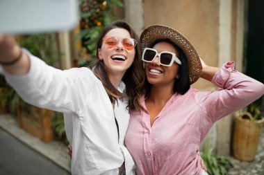 Şapkalı ve güneş gözlüklü mutlu Afro-Amerikan ve Avrupalı kadınlar akıllı telefondan selfie çekiyorlar. Birlikte şehir dışına çıkmanın tadını çıkarıyorlar. Tatil gezisi blogu, macera ve yaşam tarzı için turizm ve uygulama