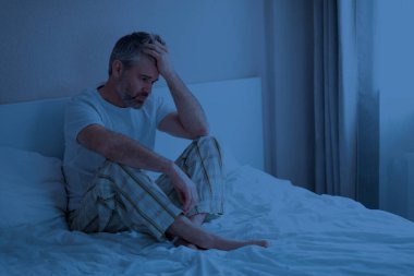 Uyku bozuklukları. Stresli, düşünceli, orta yaşlı, düşünceli, yakışıklı, pijama giyen, gece yatağa oturmuş, kafaya dokunmuş, kötü rüyadan uyanmış, kabuslardan muzdarip, ev içi, fotokopi odası.