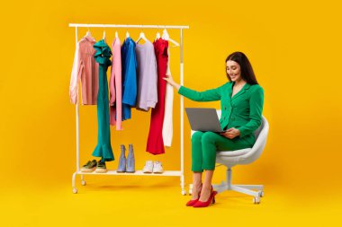 İnternetten alışveriş. Diz üstü bilgisayar kullanan ve internetten kıyafet alan şık genç bir bayan sarı stüdyo arka planında giysi korkuluğunun yanında oturuyor.