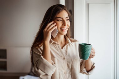 Mutlu, genç, güzel Asyalı kadın telefonla arıyor, konuşuyor, boş zamanlarında bir fincan içkinin keyfini çıkarıyor, modern oda içinde bir hafta sonu. Dedikodu, iyi haber, satış ve kahve molası, iletişim.