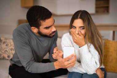 Üzgün, kızgın, bin yıllık Arap adam Avrupalı bir kadına iç odada akıllı telefon gösteriyor, kapat. Tartışma, sosyal ağların uygulaması sohbet ve blog, alet bağımlılığı, online ihanet ve ilişkiler sorunları