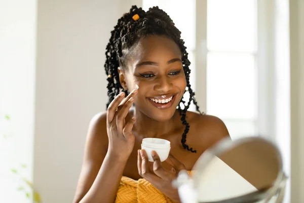 シャワー後の顔のクリームを使用して魅力的な黒人女性 毎日のスキンケアルーチンを作ります 自宅で寝室に座って 鏡と笑顔を見て 無料のスペース — ストック写真