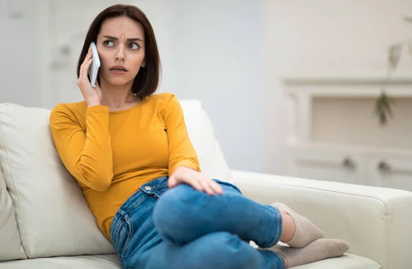 stock image Annoyed beautiful brunette young woman in comfy casual outwear sitting on couch with legs up, having phone conversation with friend or lover at home, have fight, looking aside, copy space