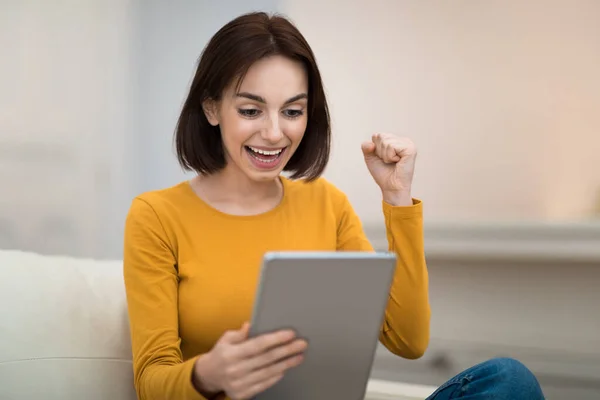 感情的に幸せなかなり若い女性でカジュアルで現代的なデジタルタブレットで彼女の手ジェスチャーと笑顔 成功を祝う パッド上のビデオゲームをプレイ ホームインテリア コピースペース — ストック写真