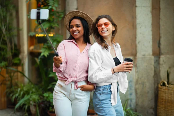 Cheerful young diverse women in hat and sunglasses make selfie on smartphone, enjoy trip in city, drink coffee takeaway outdoor. Vacation tourism and app for travel blog, photo for social networks