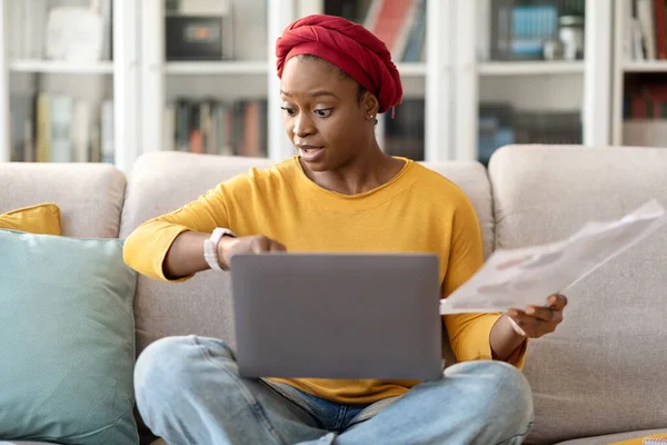 Time management concept. Worried distressed young black woman in casual entrepreneur check smart watch, sitting on couch with laptop and papers in hand, working from home, getting late for meeting