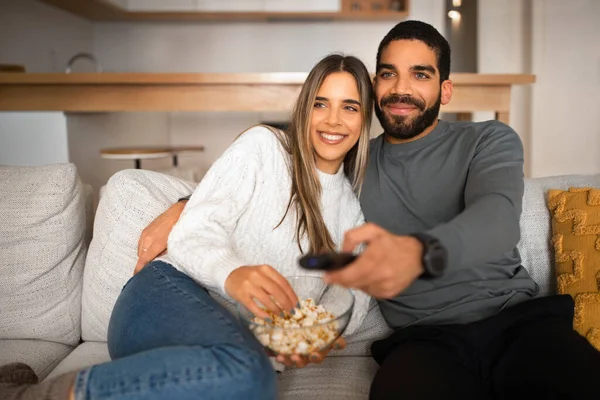Smiling millennial arabic guy with beard and remote control hugs european lady, watch TV, eat popcorn, enjoy movie together on sofa in living room. Snacks in free time, entertainment and fun at home