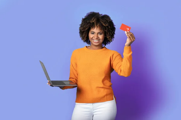 Easy banking, online shopping. Smiling cheerful beautiful young african american woman in casual holding red bank credit card and modern pc laptop on purple background, copy space