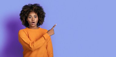 Şaşırtıcı duygusal, güzel kıvırcık, turuncu elbiseli siyah kadın reklam için fotokopi alanını işaret ediyor ve gözleri ve ağzı açık, mor arkaplan, panorama