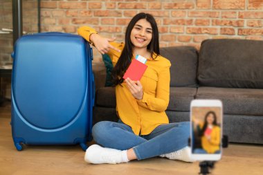 Mutlu İspanyol kadın seyahat blogcusu cep telefonuyla sosyal medya için video çekiyor, pasaportunu biletle gösteriyor, seyahat planları hakkında konuşuyor, vlog 'u için canlı yayın içeriği hakkında konuşuyor