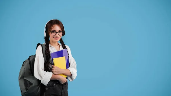 带着辫子和带着背包的无线耳机的眼镜的快乐的欧洲少女学生拿着用蓝色工作室背景 全景隔离的笔记本 现代学习 知识和教育 — 图库照片