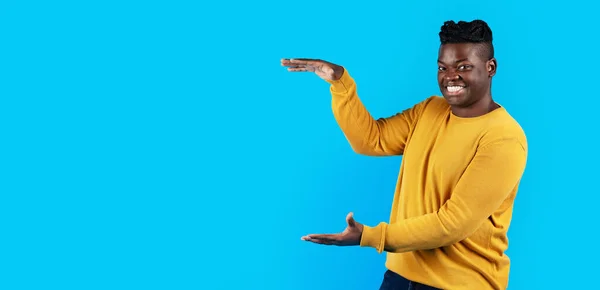 Smiling African American Man Measuring Invisible Object With His Hands, Happy Black Male Indicating Free Space For Item Advertisement, Holding Something In Arms While Standing Over Blue Background