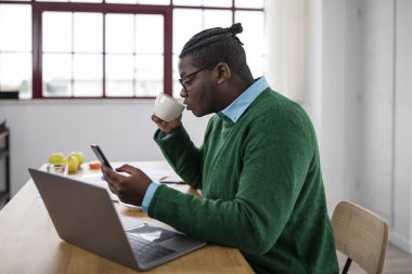 Siyahi iş adamı cep telefonu kullanıyor ve laptopta çalışıyor. Ofiste oturup kahve içiyor. İş yerindeki internet telefonuyla mesajlaşan ve gezen adam. İş Uygulaması