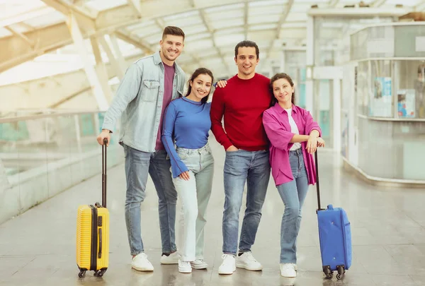 現代の空港の屋内でカメラに微笑む旅行スーツケースに立つ2人の旅行者 一緒に休暇に行く4人の若者のグループ フルレングスショット — ストック写真