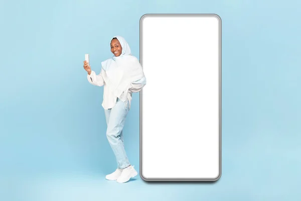 幸せなアフリカ系アメリカ人女性でヒジャーブは空の白い画面とモバイルデバイスを使用して大きな携帯電話の近くに立って ウェブサイトやアプリのデザインのためのモックアップ 青の背景 — ストック写真