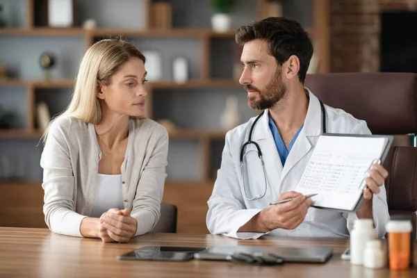 Male Therapist Talking To Female Patient During Appointment In Office, Doctor Man In White Coat Consulting Young Woman, Showing Clipboard With Test Results And Explaining Medical Therapy Process