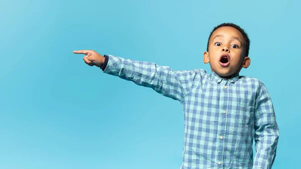 Look here. Surprised black boy pointing aside at free copy space and looking at camera with open mouth over blue background. Emotional male child showing place for ad