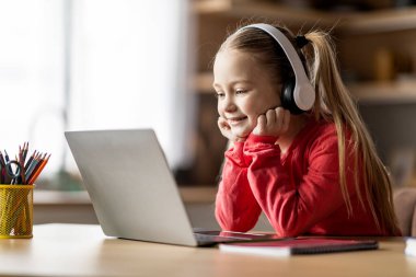 Evde dizüstü bilgisayarla kablosuz kulaklık takan, bilgisayar ekranına bakan, gülümseyen, internetten ders seyreden, uzaktan eğitimin keyfini çıkaran, boş alan arayan küçük tatlı bir kız.