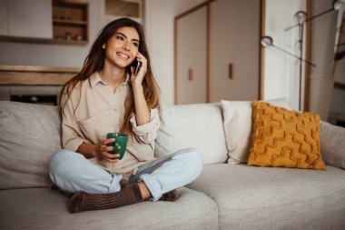 Neşeli Asyalı genç bayan telefonla arıyor, konuşuyor, kanepeye oturuyor, sıcak oturma odasında boş vakitlerinde bir bardak içkinin tadını çıkarıyor. Dedikodu, harika haberler, satış ve kahve molası, günaydın.