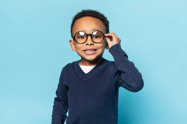 穿着校服的神经质的非洲裔美国男孩摸着眼镜 看着镜头 微笑着 在蓝色的画室背景下调整圆形眼镜 — 图库照片