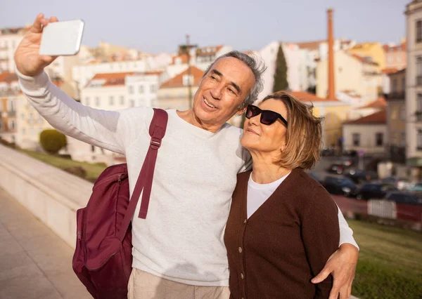 Vacation Travel. Happy Senior Tourists Couple Making Selfie On Phone Hugging Standing With Backpack Outdoors. Spouses Traveling Walking In Lisbon Enjoying City Tour. Tourism And Sightseeing