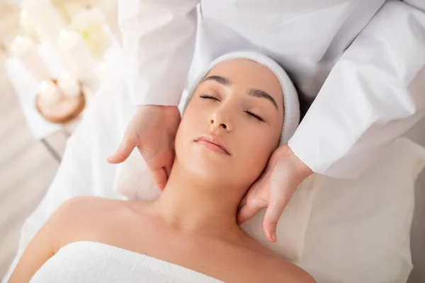 Young indian female enjoying therapeutic neck massage in spa, closeup shot of beautician doctor doing beauty treatments to calm woman lying on massage table in wellness center, above view
