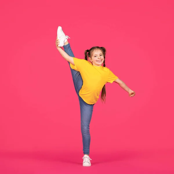 Cute little gymnast girl raising her leg in twine while posing over pink studio background, cheerful preteen female child in casual clothes making gymnastics, doing stretching exercises, full length