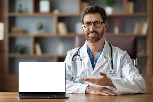 Great Website. Friendly Doctor Man Pointing At Laptop With Blank White Screen While Sitting At Desk In Clinic Office, Smiling Male Therapist Showing Free Place For Medical Ad Design, Mockup