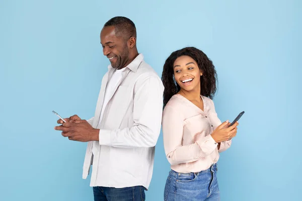 不信の概念 好奇心旺盛な黒人女性は夫のスマートフォンを見て 男と女を笑って ガジェットを使っておしゃべりし カップルは青いスタジオの背景に立って — ストック写真