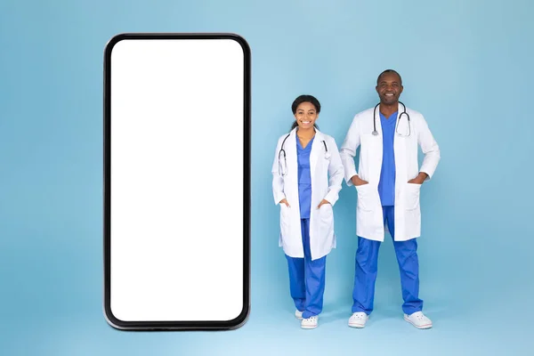 Black male and female doctors standing near huge smartphone with blank screen, posing over blue background, mockup. Professional physicians advertising medical mobile app