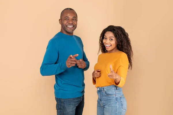 Positive black couple pointing at camera with fingers and smiling, african american spouses indicating somebody, standing together over beige studio background, copy space