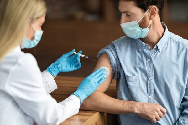 戴手套的女医生 给戴医疗面具的男病人注射疫苗 把注射器插到他的胳膊上 在现代诊所给年轻人注射预防考拉病毒的疫苗 — 图库照片