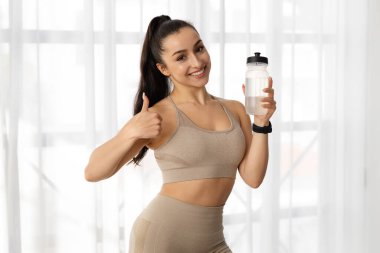 Spor giyim kuşamında seksi ve çekici genç bir kadın spor salonunda ya da evde pencerenin önünde duruyor, elinde bir şişe su tutuyor, başparmağını kaldırıyor ve kameraya gülümsüyor, idman yaparken su kaybediyor.